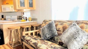 A cozy living area with a sofa adorned with a nature-themed print, two fluffy grey cushions, and a wooden coffee table. A kitchen area with a microwave and wooden cabinets is visible in the background. - 4 Cabins/Mountain Views /Jacuzzi/Wi-Fi/Large Groups (Whittier)