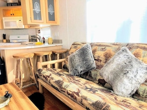 Ein gemütlicher Wohnbereich mit einer mit einem Naturmotiv versehenen Couch, zwei flauschigen grauen Kissen und einem Holztisch. Im Hintergrund ist eine Küchenzeile mit einer Mikrowelle und Holzschränken zu sehen.