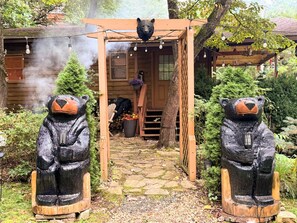 Une cabane en bois, ornée d’une statue d’ours à l’entrée, avec un chemin de pierres et une cheminée d’où s’échappe de la fumée.
