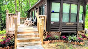 A wooden cabin with a wooden staircase leading to a deck, surrounded by a garden with various plants and flowers.