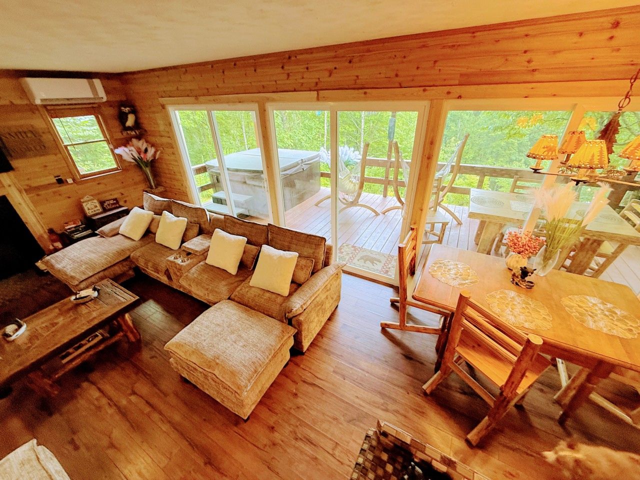 A cozy cabin interior with a large wooden sofa, a dining table with chairs, and a large window offering a view of the outdoors.