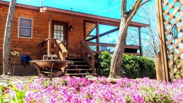 A wooden cabin with a porch, a wheelbarrow, and a garden with pink flowers.