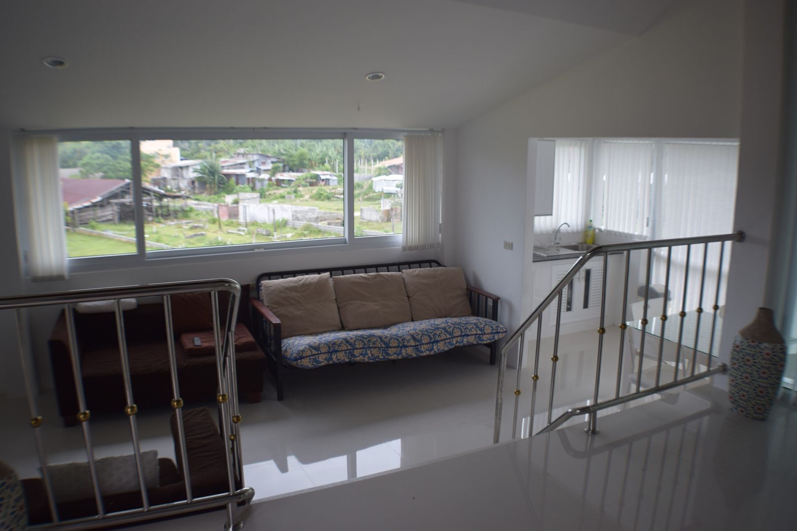 A spacious room with a high ceiling, a large window offering a view of a residential area, a sofa with patterned cushions, and a metal staircase railing.