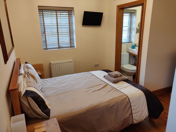 A bedroom with a bed, a window, a radiator, a TV, and a bathroom visible through an open door.