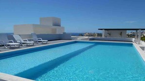 A rectangular swimming pool with clear blue water, surrounded by white lounge chairs and a modern building structure. - Beautiful Condo 300 Meters To Medano Beach (Cabo San Lucas)