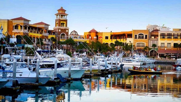 A marina with boats docked, a tower, and buildings with balconies. - Beautiful Condo 300 Meters To Medano Beach (Cabo San Lucas)