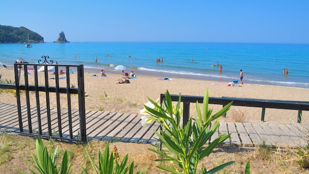 Ein Strand mit einer Holzpromenade, die zum Ufer führt, klarem, blauen Wasser und einer in der Ferne liegenden Felsformation.