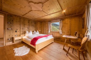 A wooden cabin bedroom with a bed, bedside table, chair, and a window with a view of the outdoors. - Charming Apartment “Panorama Raffeinhof” with Lake View, Balcony, Terrace & Garden (St. Walburg, Südtirol)
