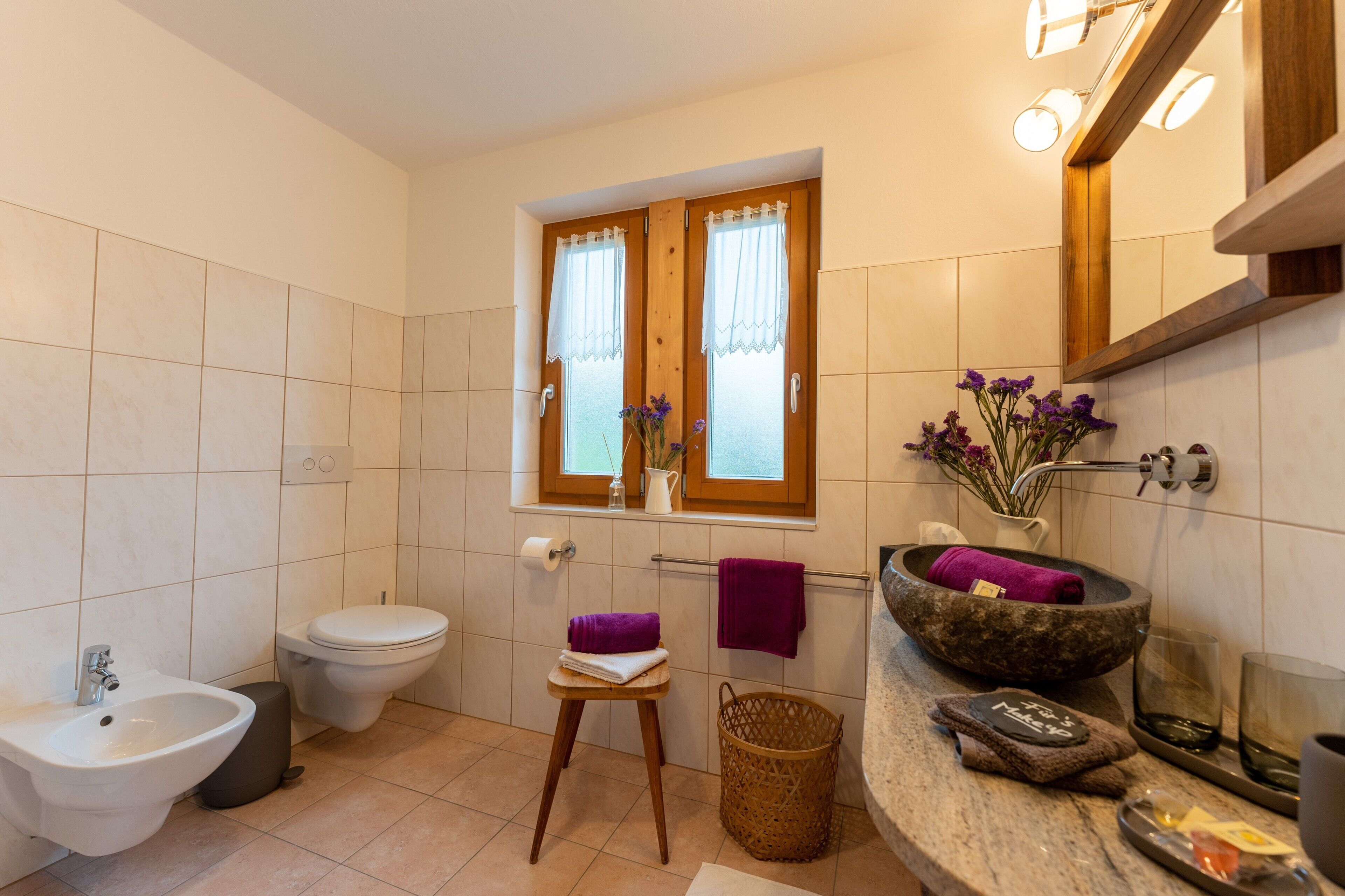 Un bagno con bidet, WC, mobile lavabo e finestra con tende.