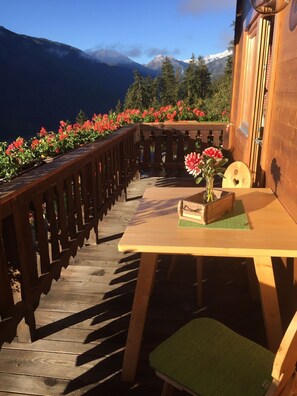 A wooden deck with a table and chair, a vase with flowers, and a view of mountains. - Charming Apartment “Panorama Raffeinhof” with Lake View, Balcony, Terrace & Garden (St. Walburg, Südtirol)