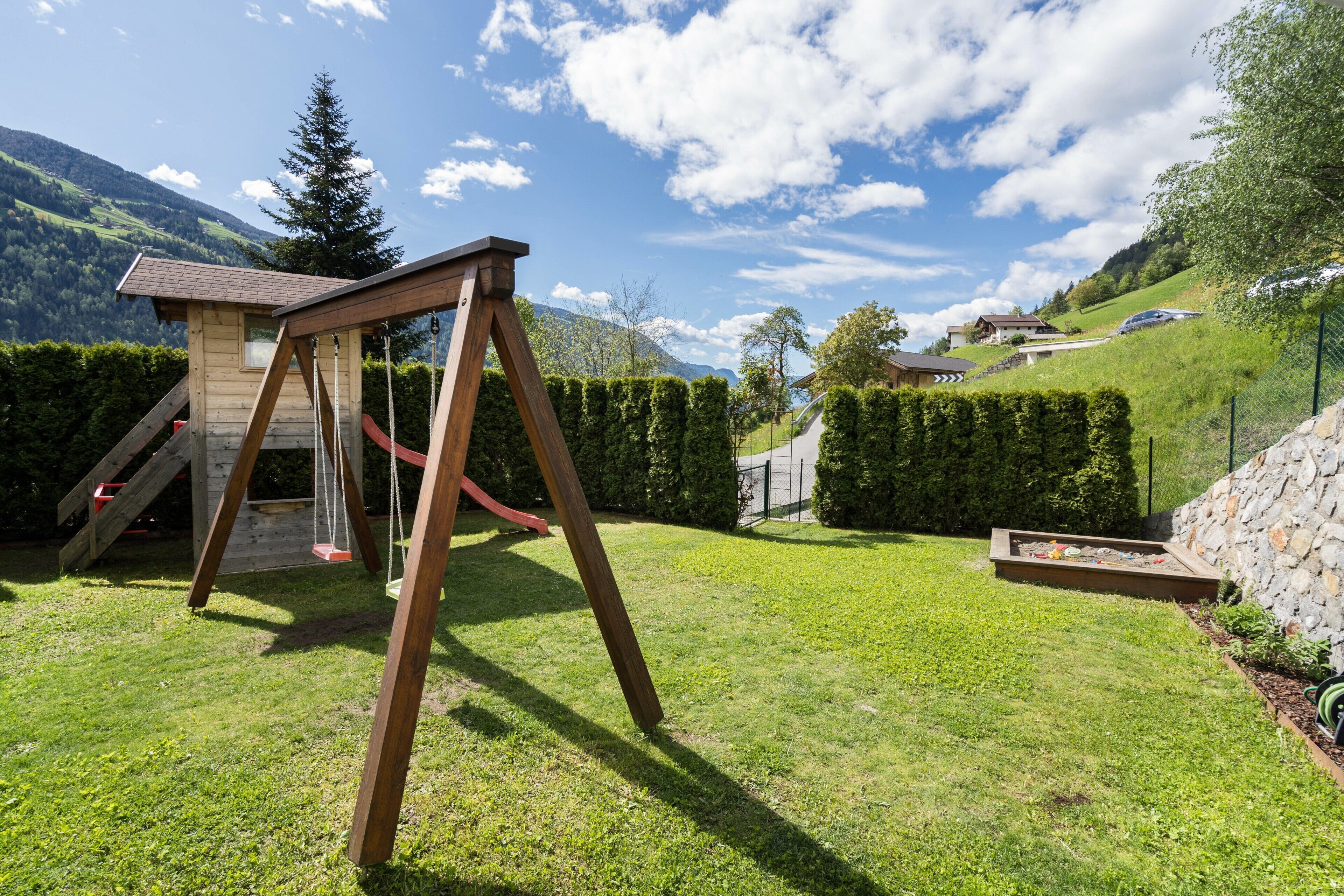 A wooden swing set in a grassy backyard with a stone wall and a view of a mountain.