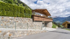 A stone-walled building with a wooden balcony, a paved road, and a mountainous landscape in the background.