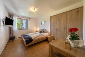 A bedroom with a bed, a TV, a window, a wooden wardrobe, and a table with a vase of flowers.