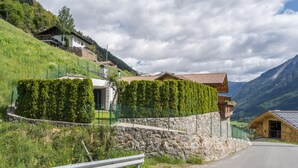 A hillside house with a stone wall, a wooden building with a sloped roof, and a road with a metal guardrail.