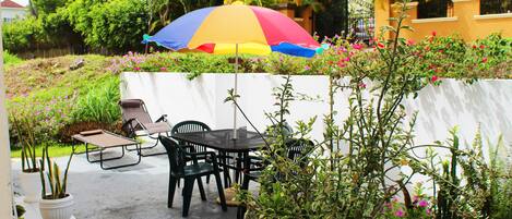 Un patio al aire libre con una mesa redonda y sillas, rodeado de macetas con plantas y un paraguas colorido.
