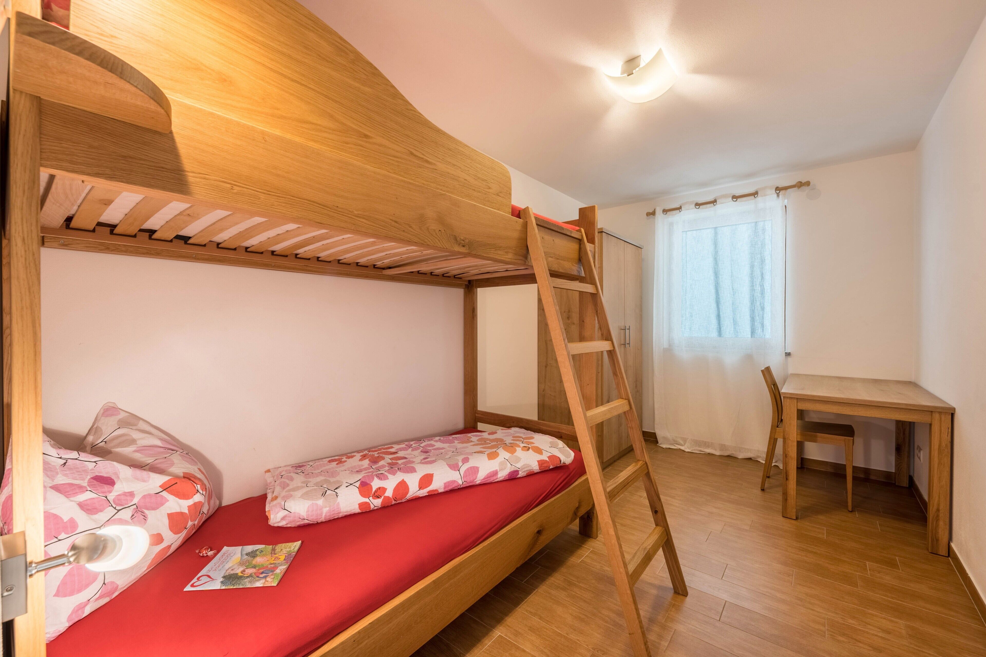 Een stapelbed met een ladder, een klein bureau en een stoel in een kamer met houten vloer en witte wanden.