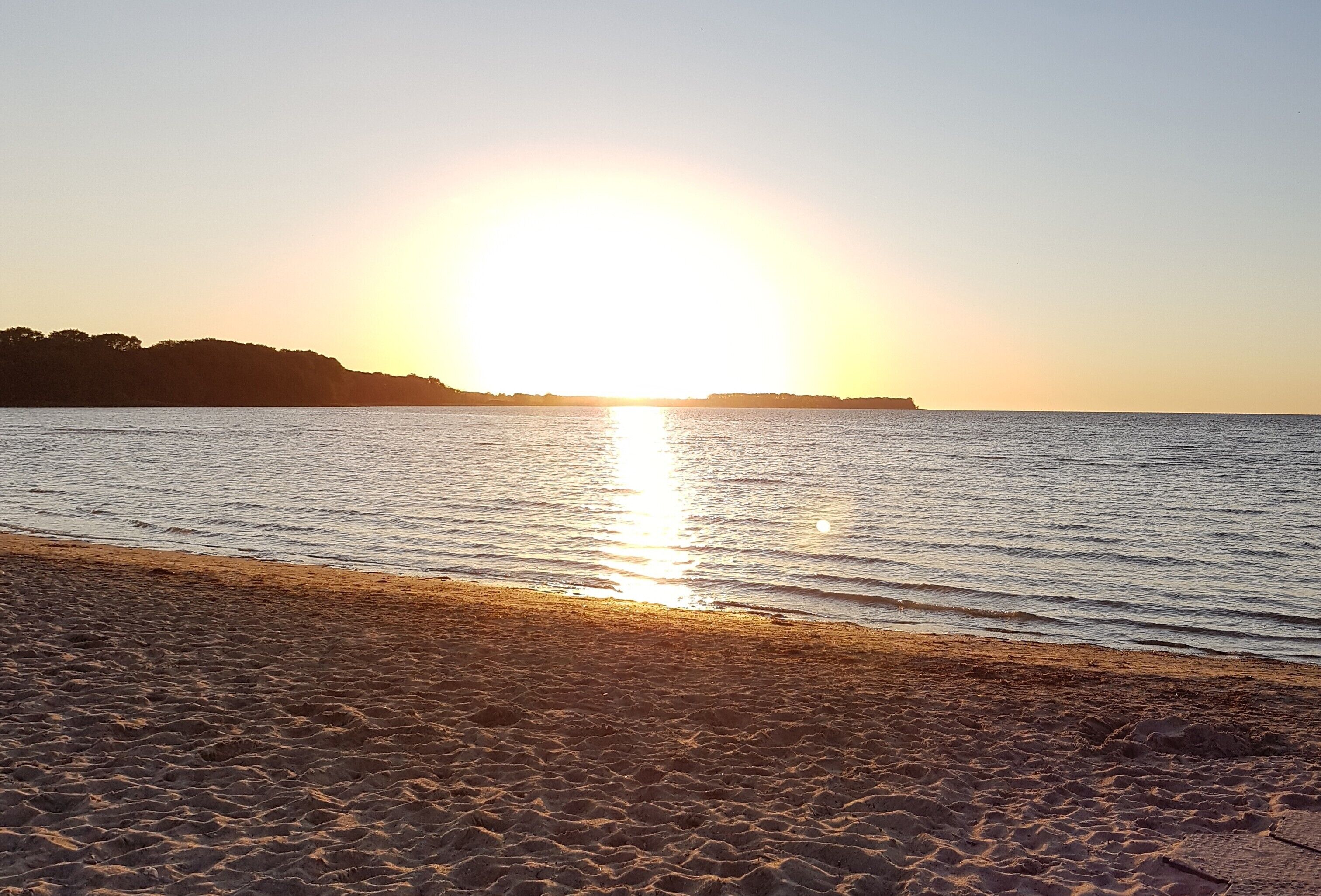 A beach with a sunset over the water, sandy shore, and distant land.