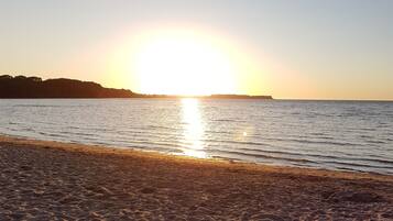 A beach with a sunset over the water, sandy shore, and distant land.