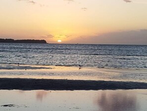 Een strand met een zonsondergang boven de oceaan, kalm water en een verre kustlijn.