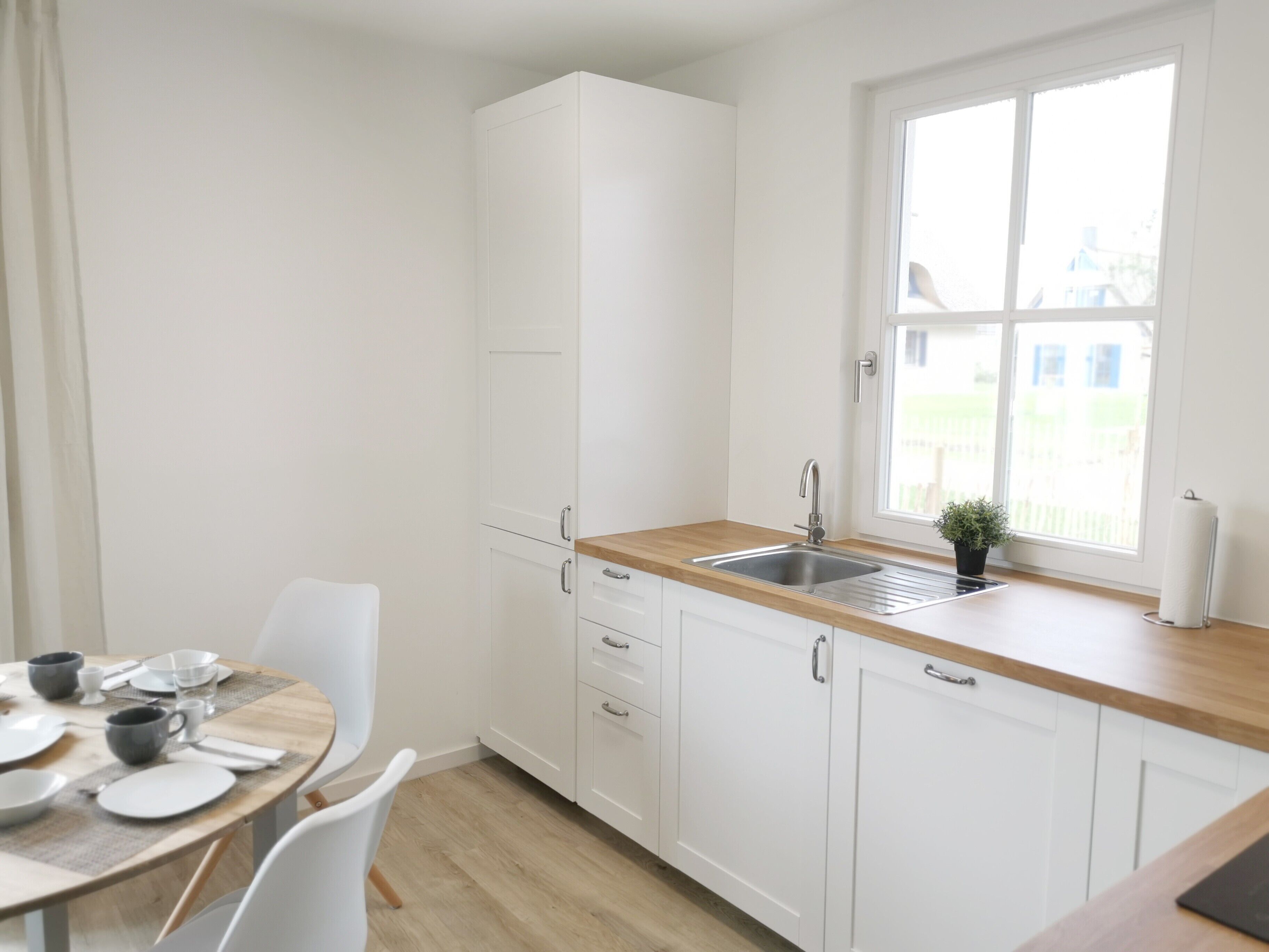 Une cuisine moderne avec des meubles blancs, un plan de travail en bois et une fenêtre donnant sur une maison.