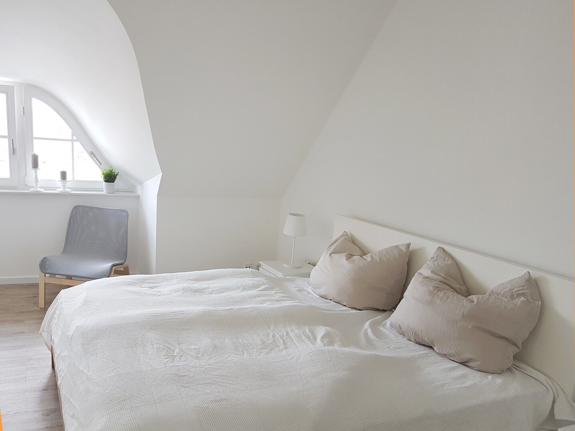 Un dormitorio con cama, almohadas, una silla, una mesita de noche y una ventana.