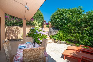 Eine Terrasse im Freien mit Korbmöbeln, einem Tisch gedeckt mit Tellern und einer Flasche, und Blick auf einen Garten mit Stufen, die zu einer Tür führen.