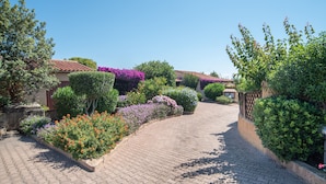 A paved pathway leading to a building with a red-tiled roof, surrounded by lush greenery and flowering plants.