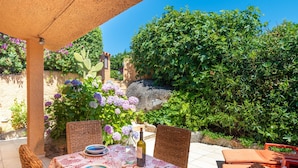 An outdoor dining area with wicker chairs, a table set with plates and a bottle, surrounded by greenery and a rock.