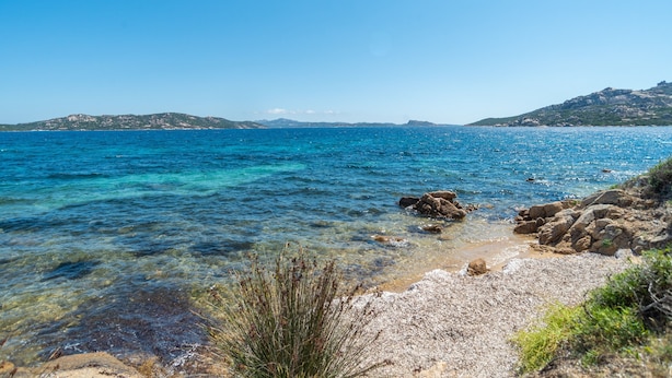 A coastal landscape with clear blue sea, rocky shore, and distant hills.