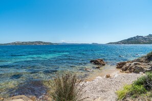 A coastal landscape with clear blue sea, rocky shore, and distant hills.