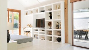 Un soggiorno moderno con una grande TV, scaffali bianchi con oggetti decorativi, un divano grigio e una porta in legno che dà sull'esterno.