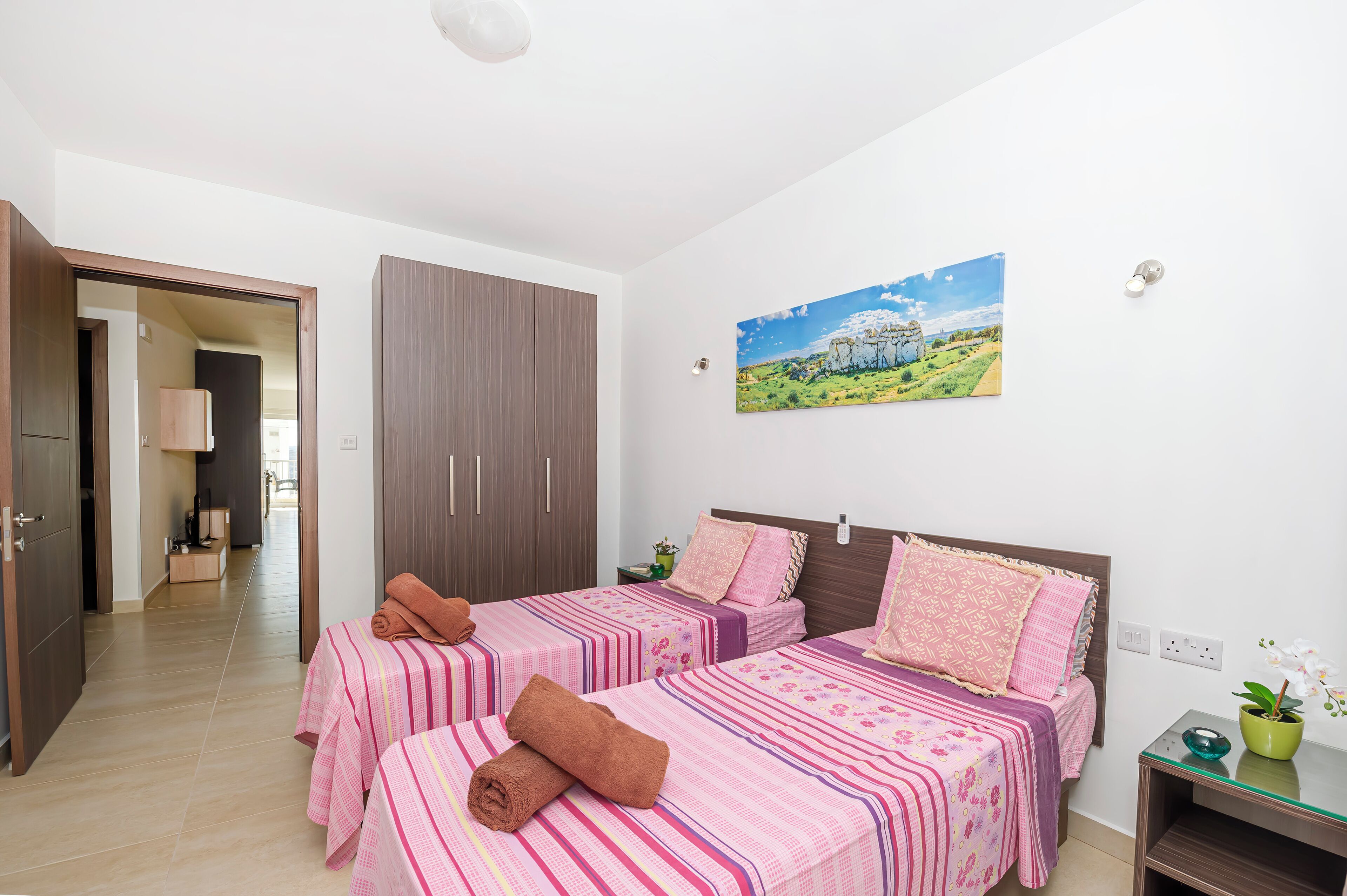 Una habitación de hotel con dos camas, un armario, un cuadro y una mesita con una planta.
