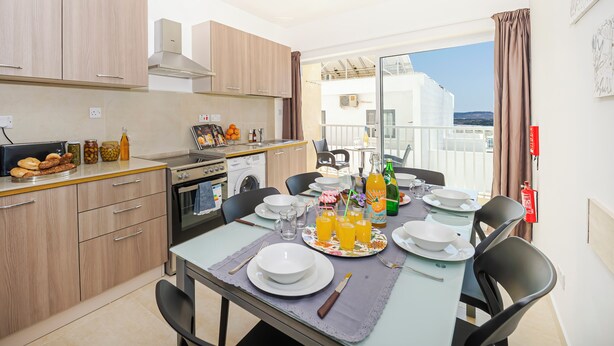 A modern kitchen with wooden cabinets, a dining table set with plates, glasses, and a bottle of orange juice. - Summer Breeze Comfort Apartment (Mellieha)