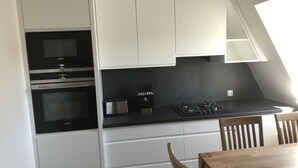 A modern kitchen with white cabinets, a black countertop, and a built-in oven. There is a wooden dining table and chairs in the foreground.