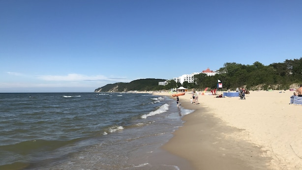A beach with a sandy shore, waves, and a few people. There are buildings in the background.