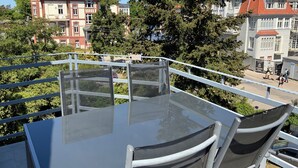 A balcony with a glass table and chairs, overlooking a cityscape with historic buildings and trees.