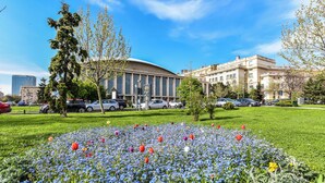 Un edificio grande con fachada curva, rodeado de un césped bien cuidado con diversas flores y árboles.