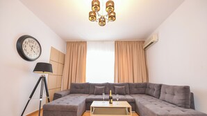 Una sala de estar moderna con un sofá seccional, una mesa de centro con una botella y vasos, una lámpara de pie, un reloj de pared y un accesorio de iluminación en el techo.