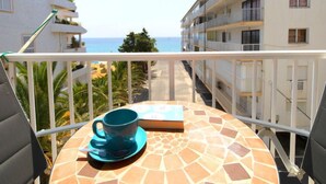 Ein Balkon mit Meerblick, ein Tisch mit Tasse und Buch sowie ein weißes Gebäude mit Balkonen.