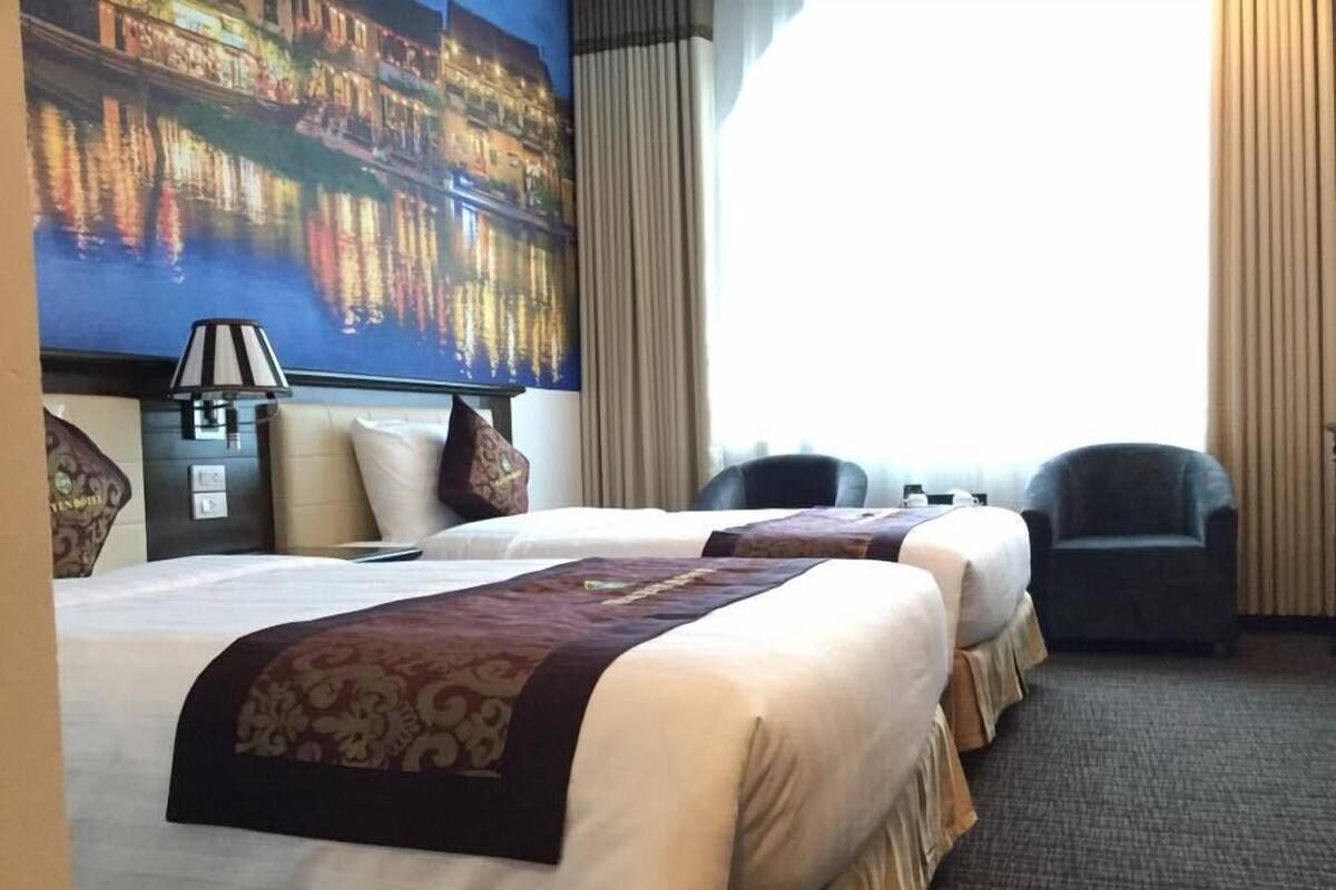 A hotel room with two beds, a large window, a lamp, and a chair.