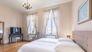 Un dormitorio con una cama, un escritorio con televisor, una silla y un ventanal con cortinas.