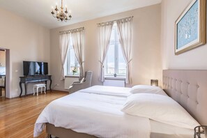Un dormitorio con una cama, un escritorio con televisor, una silla y un ventanal con cortinas.