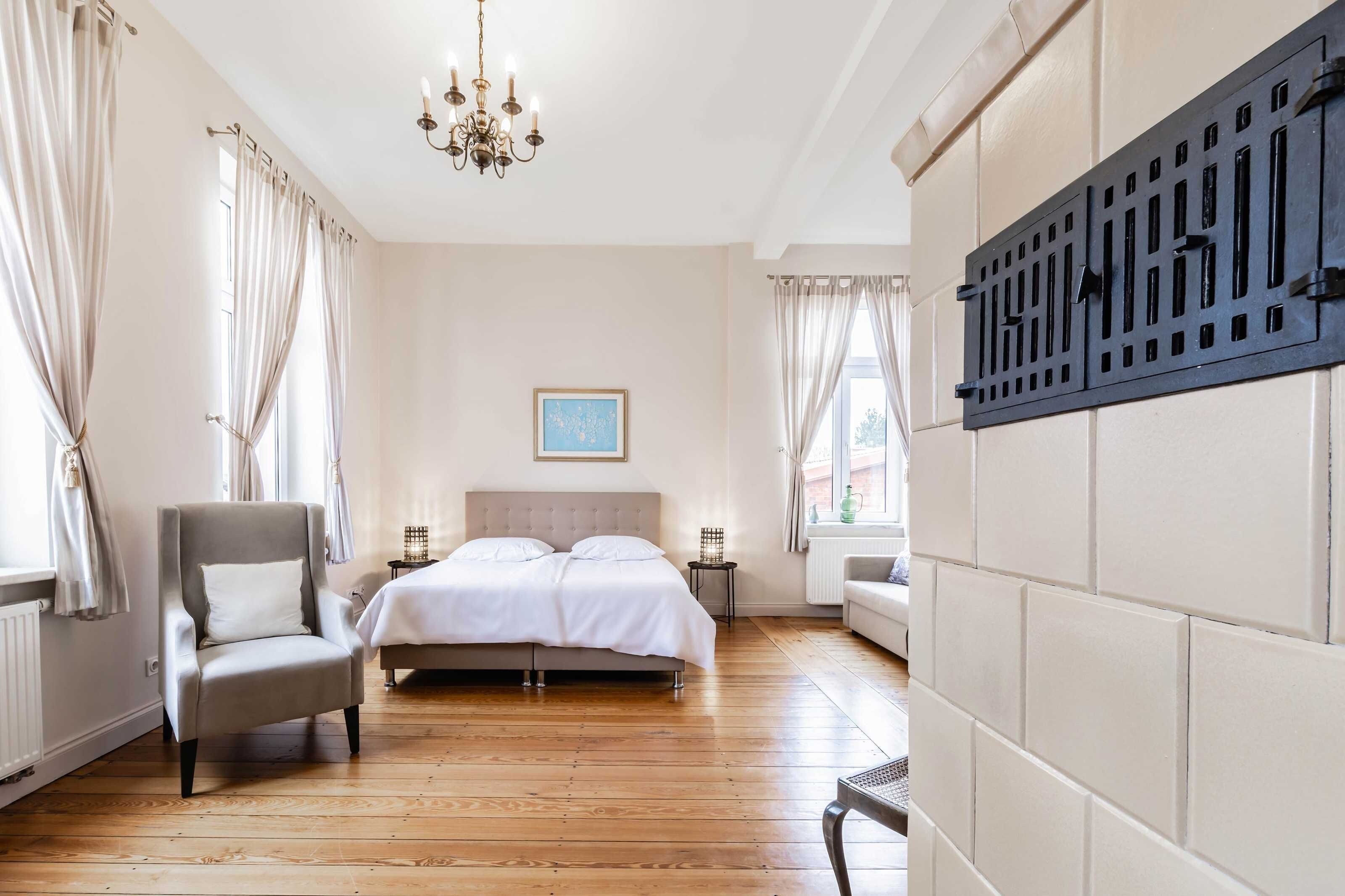 Ein Schlafzimmer mit Bett, Sessel, Nachttisch, Heizkörper und einem Fenster mit Vorhängen.