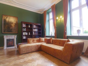 Une pièce comprenant un canapé d’angle, une cheminée, une bibliothèque et une grande fenêtre avec des rideaux.