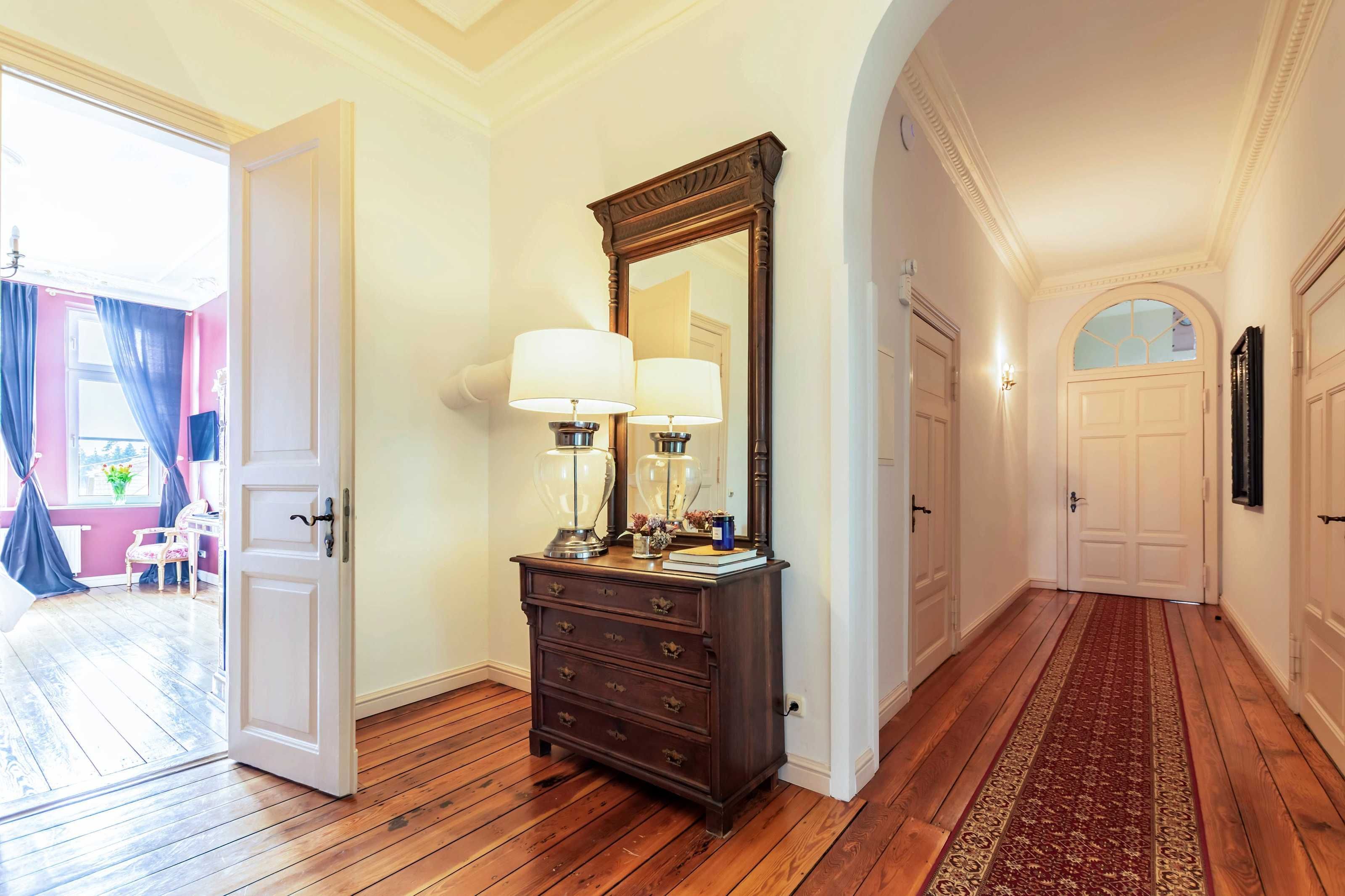 Un pasillo con suelo de madera, un aparador con lámparas, un espejo y una puerta que da a una habitación con pared rosa y una ventana con cortinas azules.
