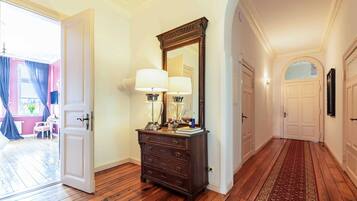 Un pasillo con suelo de madera, un aparador con lámparas, un espejo y una puerta que da a una habitación con pared rosa y una ventana con cortinas azules.