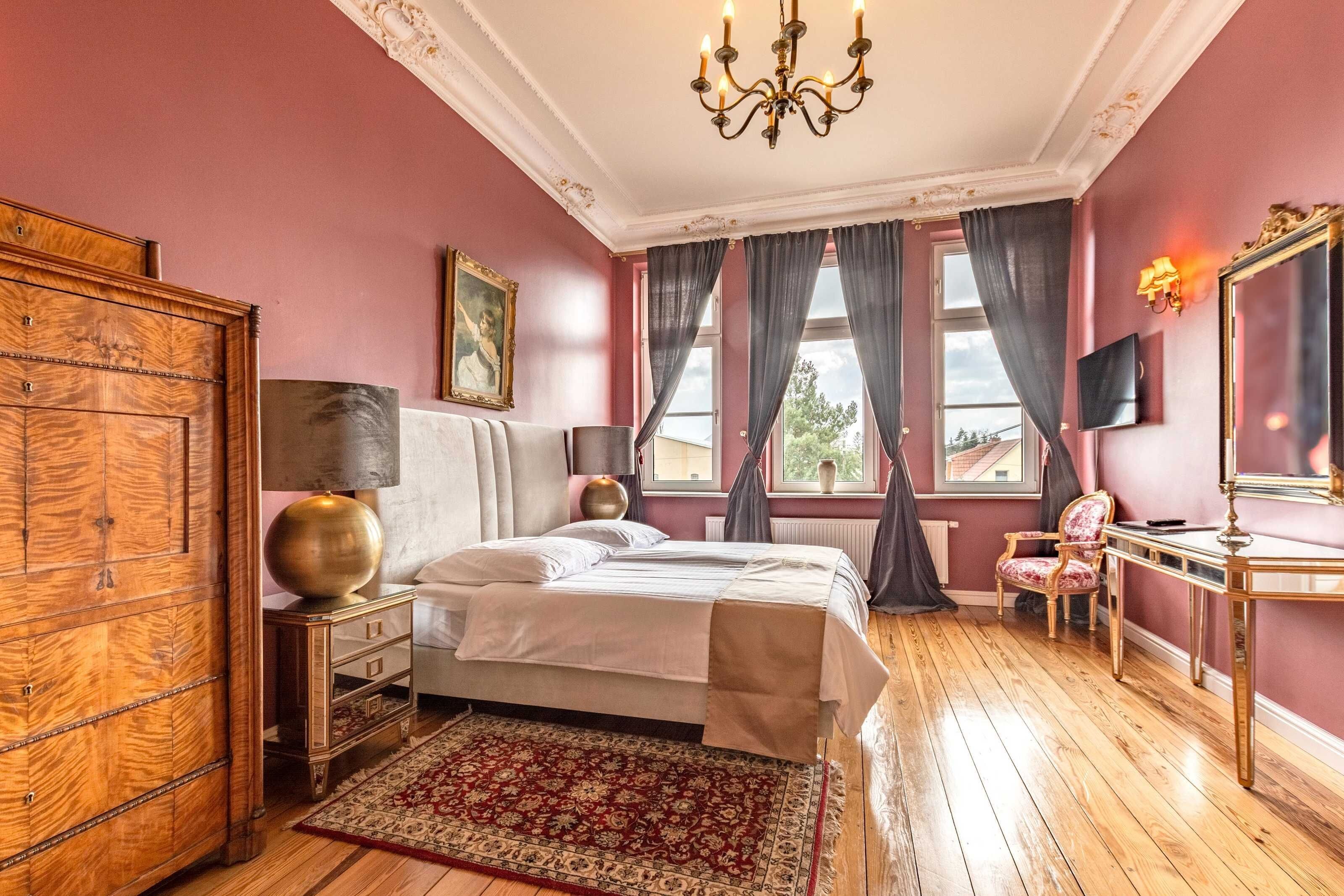 Ein Schlafzimmer mit Holzboden, einem Bett mit weißen Leinen, einem großen Spiegel, einem hölzernen Kleiderschrank und einer Kristallleuchter.