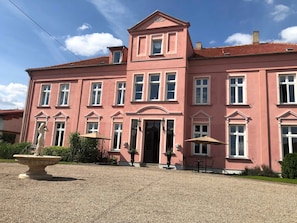 Un bâtiment rose doté de nombreuses fenêtres, d’une entrée centrale et d’un espace extérieur aménagé pour s’asseoir.
