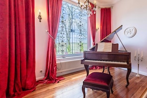 Un piano de cola, cortinas rojas, un radiador y una araña en una habitación con suelo de madera.