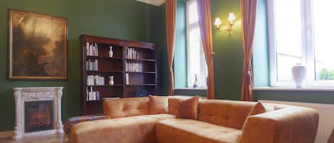 A room with a sectional sofa, a fireplace, a bookshelf, and a large window with curtains.